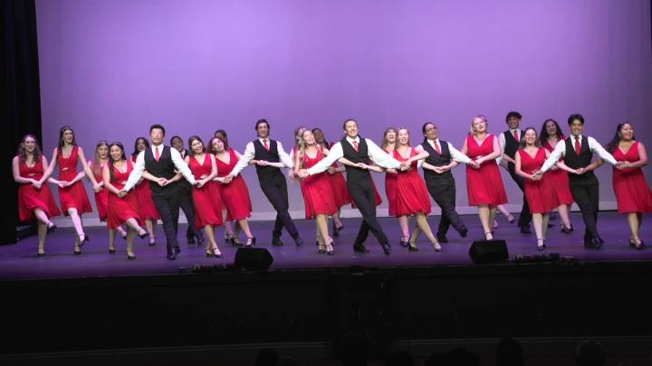 A group of dancers in unison on stage singing and dancing in red dresses and suits.