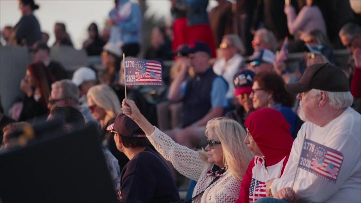 A woman in a group of people holds up a small US flag with the dates 1776 and 2026 on it.
